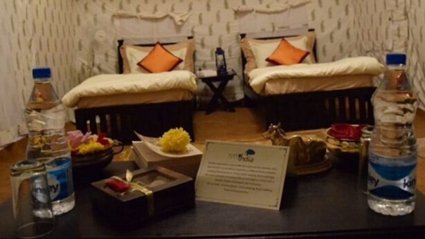 A hotel room with two beds, each with a pillow and a blanket. There are two bottles of water, a box of tissues, a small plant, and a sign with the hotel's name. - Prayag Divine Kumbh Camp - Hostel (Karchana)