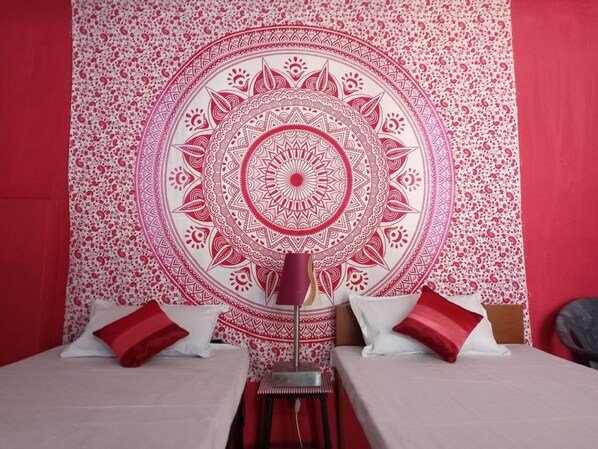 A room with a large red and white tapestry, two beds with red and white pillows, and a small table with a lamp.