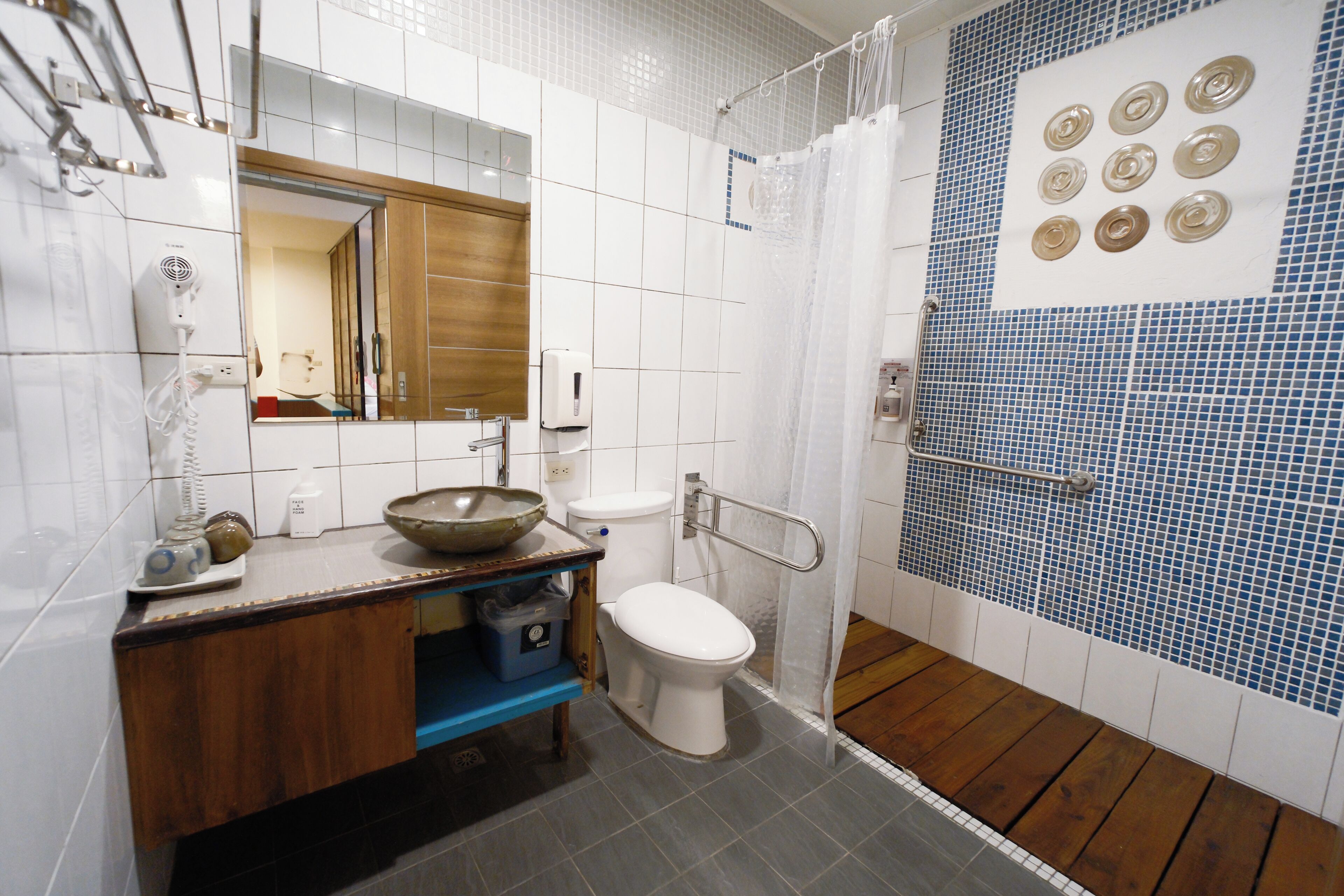 A bathroom with a white toilet, a wooden vanity, a sink with a bowl, a mirror, a hand dryer, and a shower with blue mosaic tiles.