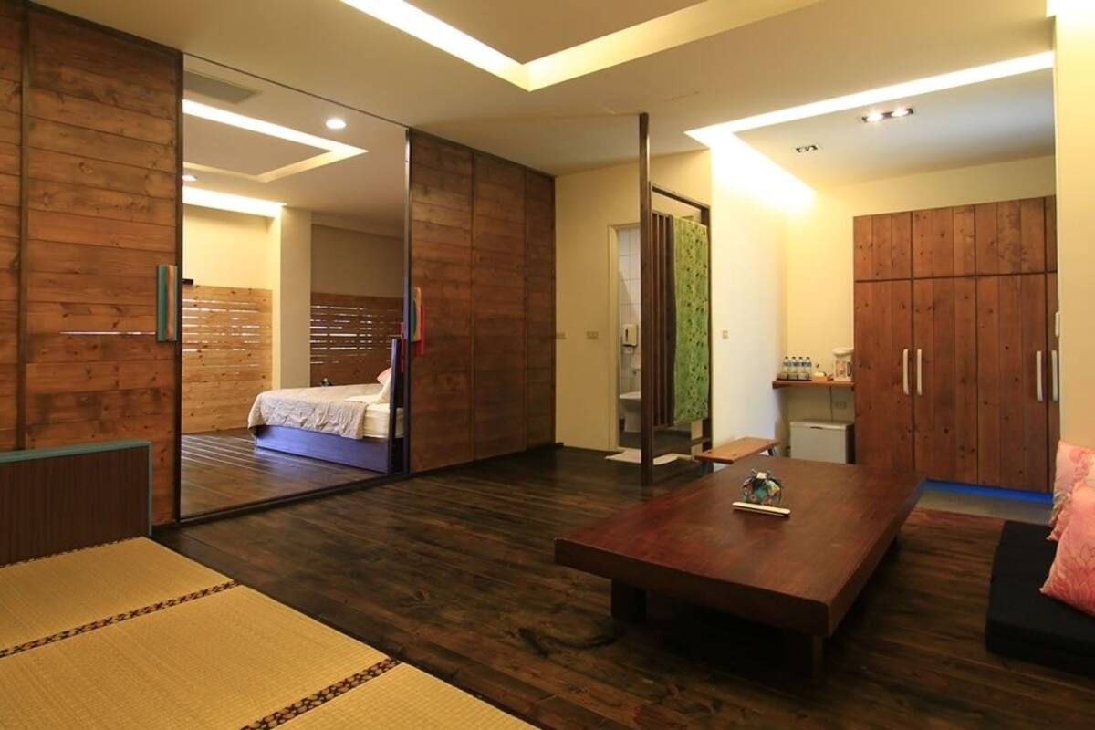 A modern hotel room with a sliding wooden partition, a bed with a white duvet, a wooden coffee table, and a small kitchenette area.
