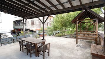 Um pátio externo com mesa e cadeiras de madeira, área de estar coberta e vista para edifícios e árvores ao fundo.