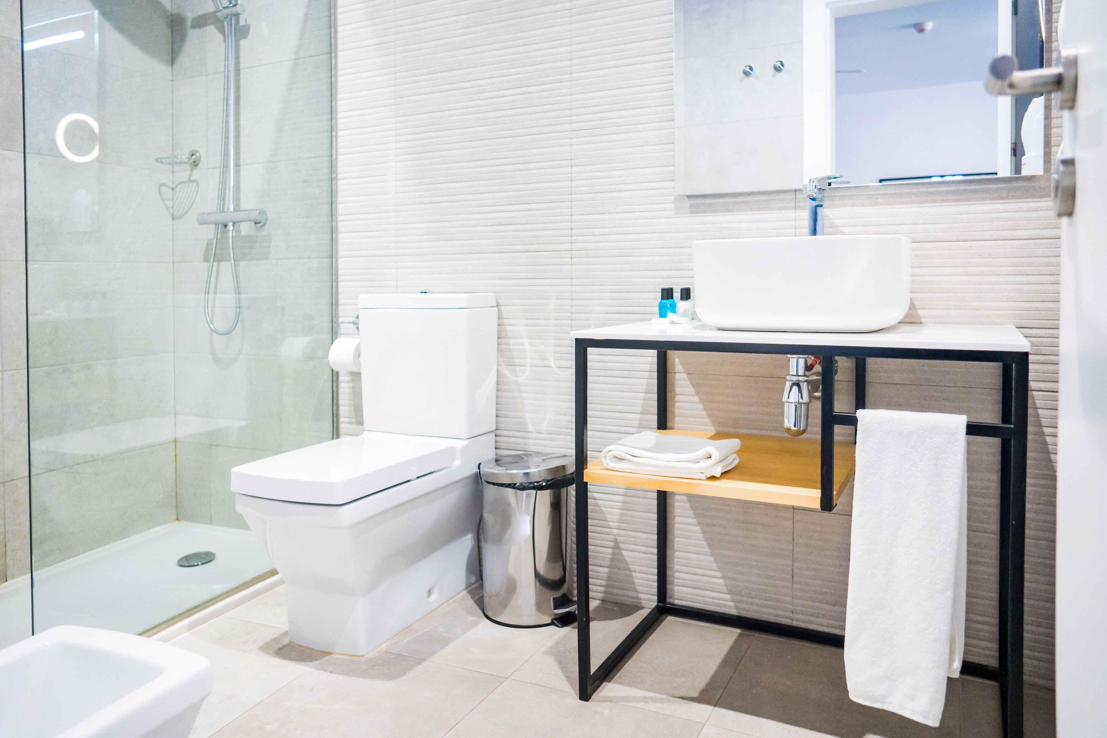 A modern bathroom with a glass shower enclosure, a white toilet, a stainless steel trash can, and a wooden vanity with a sink and towel rack.