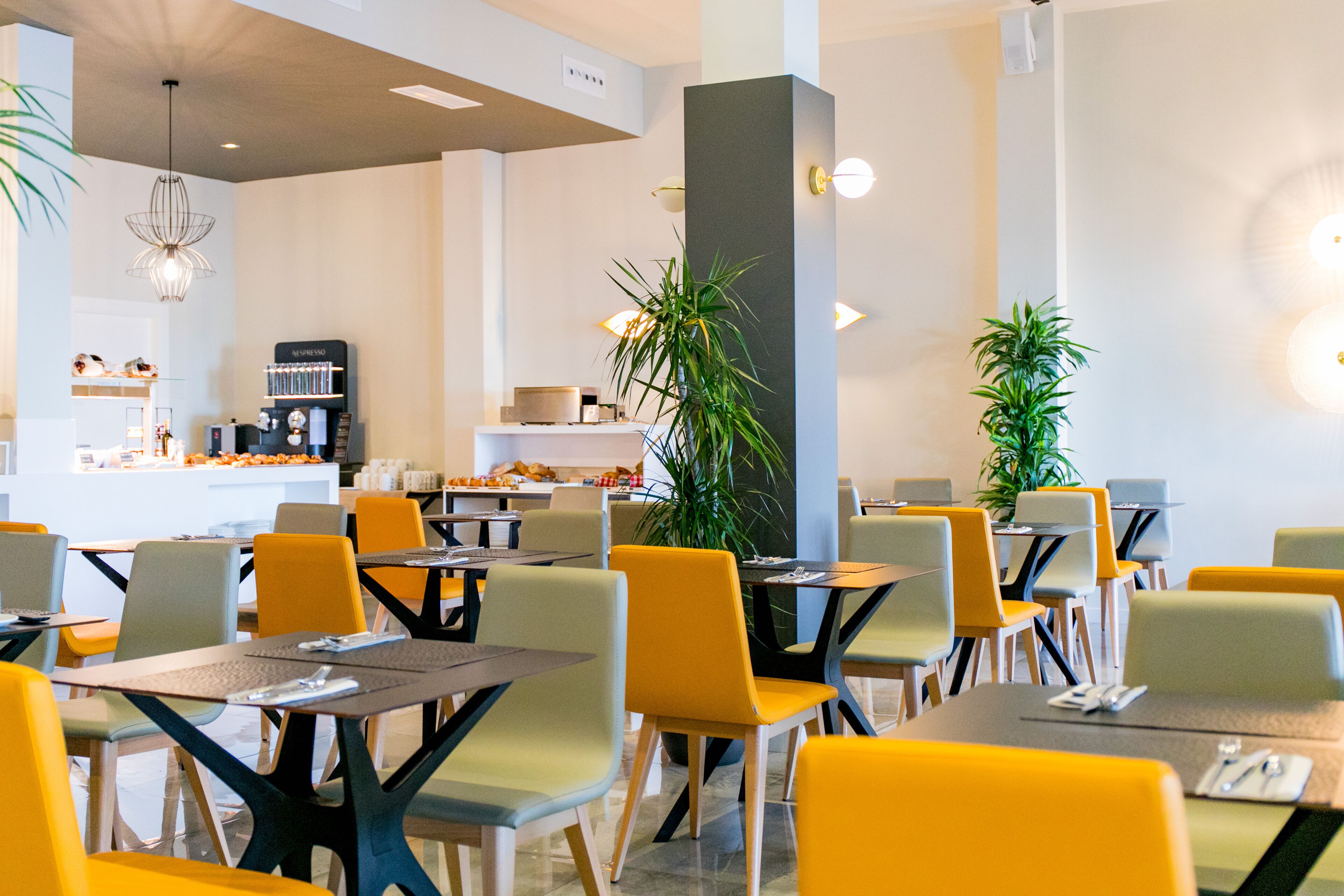 A modern dining area with yellow and grey chairs, tables, and a central plant.