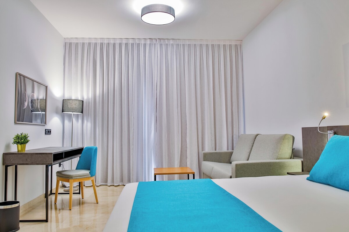 Habitación de hotel con cama, escritorio, silla, mesita, lámpara y cortina.
