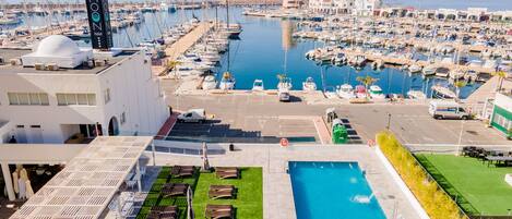 A marina with numerous boats, a swimming pool with sun loungers, and a hotel building with a sign.