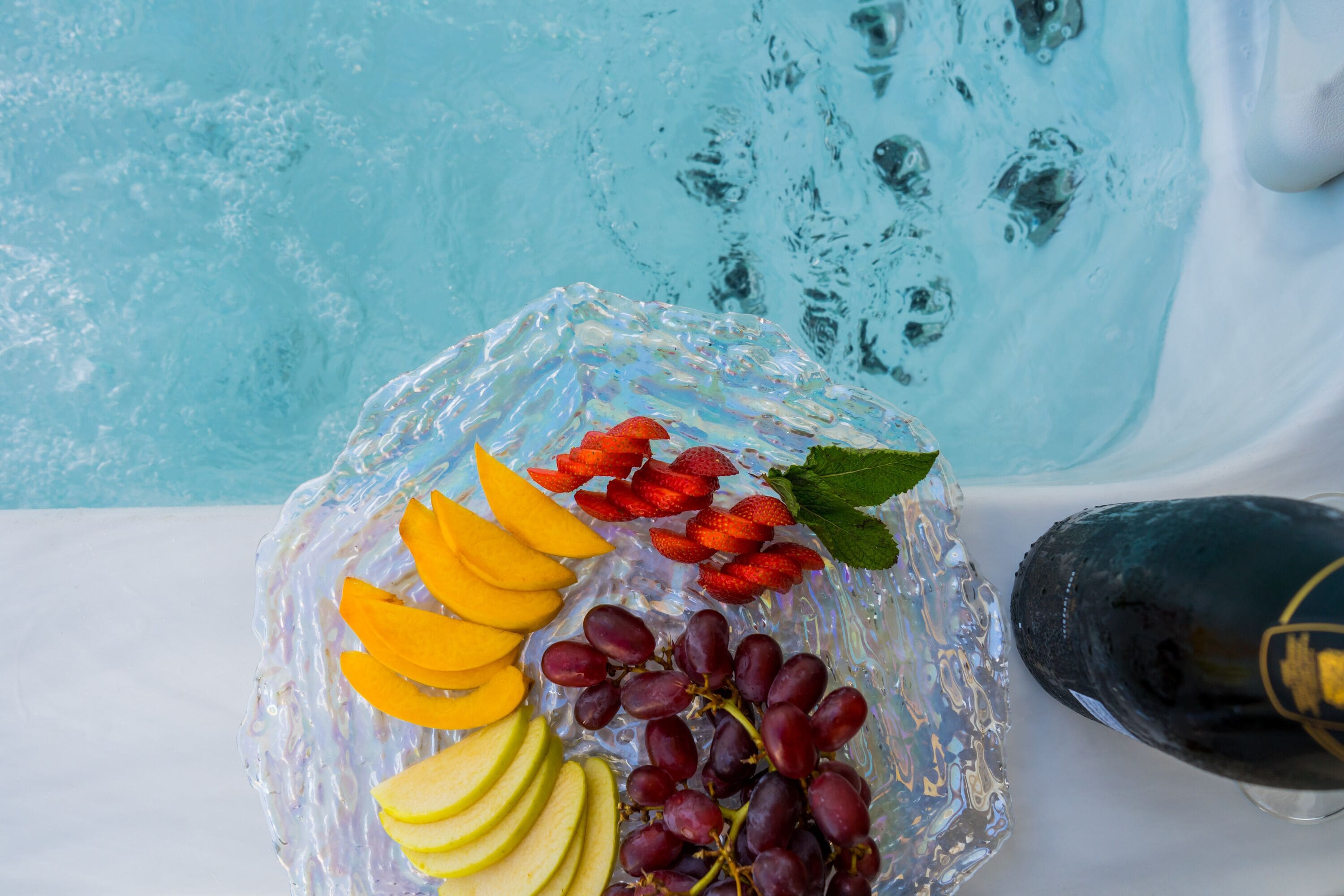 Un grand plat en verre contenant des fruits coupés en tranches et une bouteille de vin, le tout placé près d’une piscine.