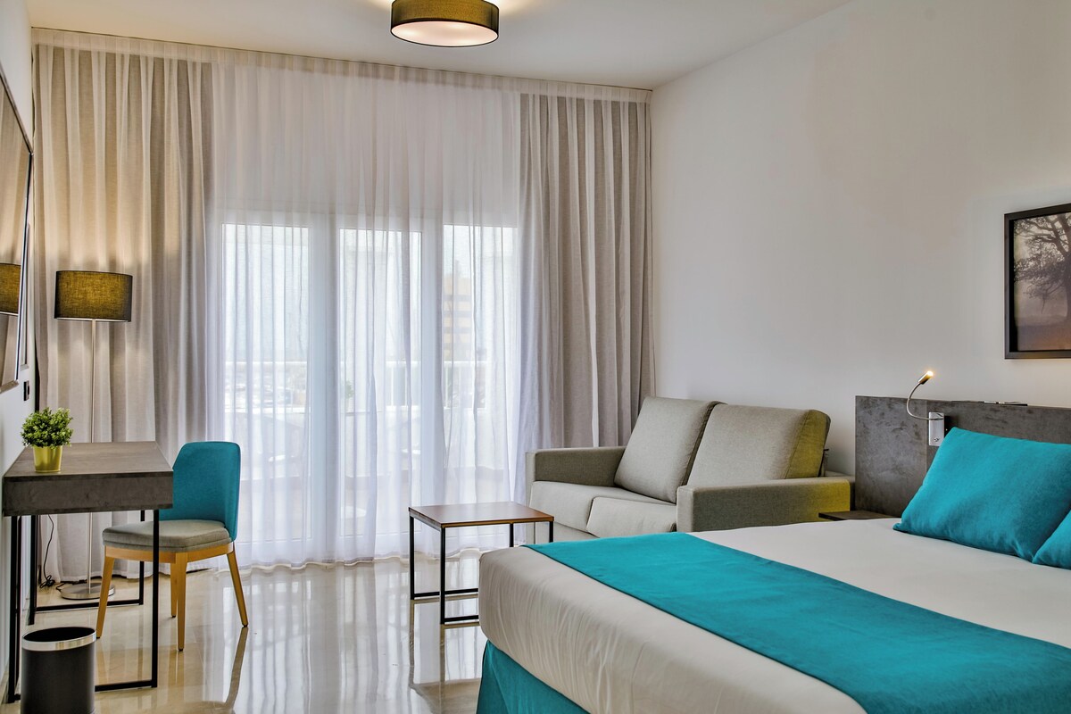 Una habitación de hotel moderna con cama, escritorio, sillón, sofá y un ventanal con cortinas translúcidas.