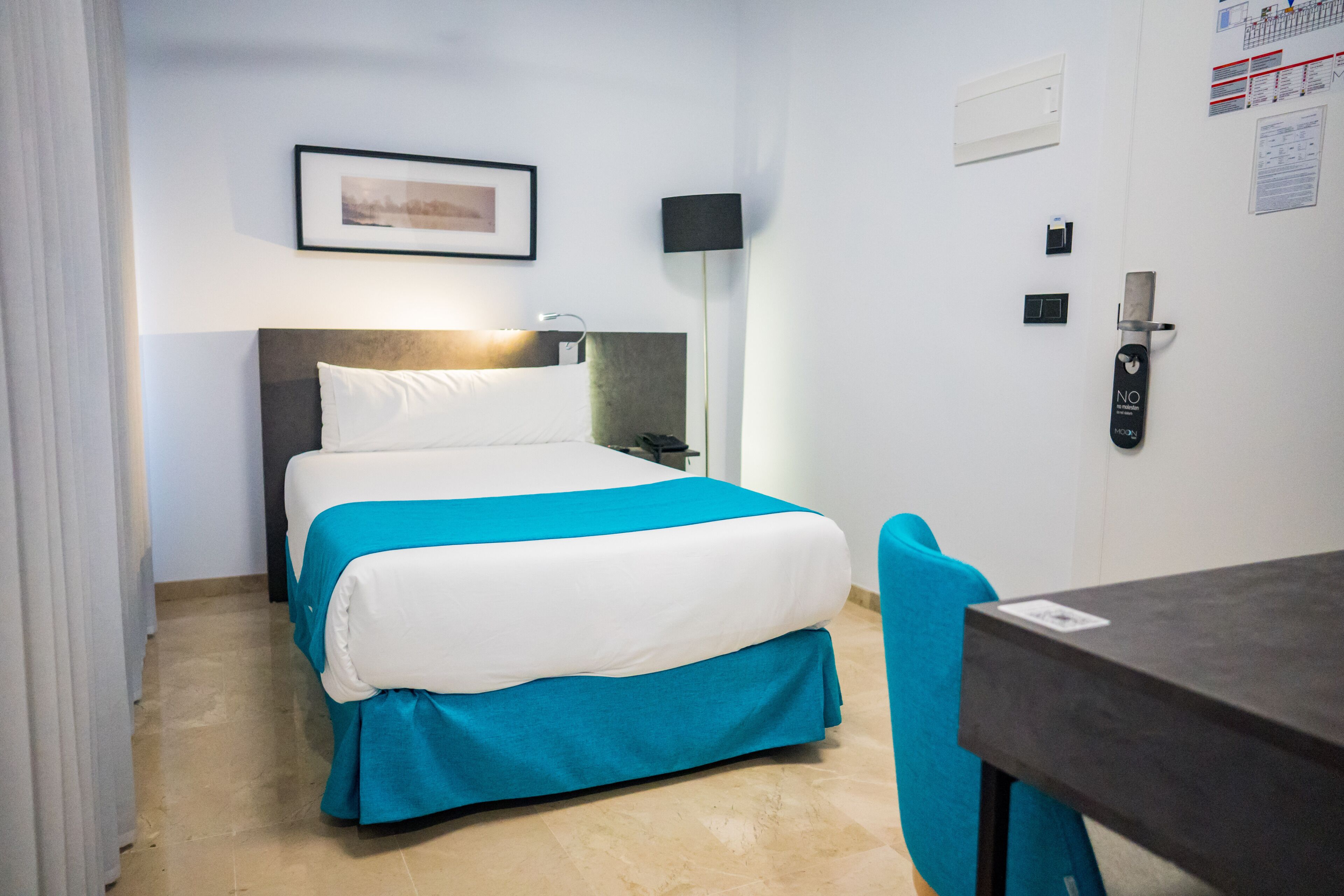 A hotel room with a bed, a desk, a chair, a wall-mounted lamp, and a door with a lock.