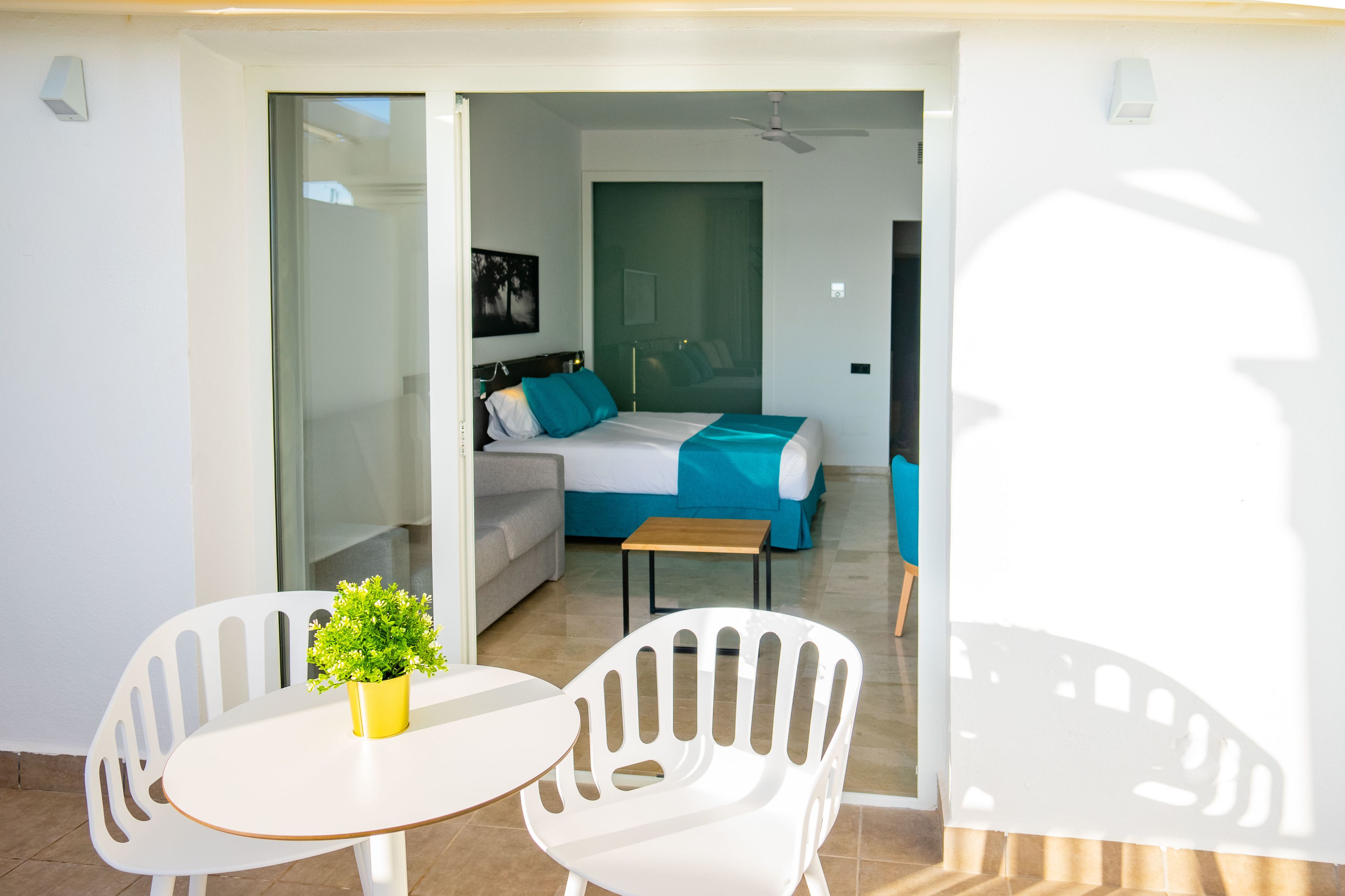 Una moderna camera d'albergo con un balcone, un letto con lenzuola blu, un divano e un tavolino con una pianta.