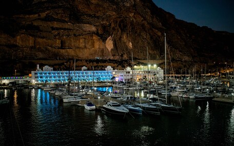 A marina at night with boats docked, a building with blue lights, and a mountain in the background.