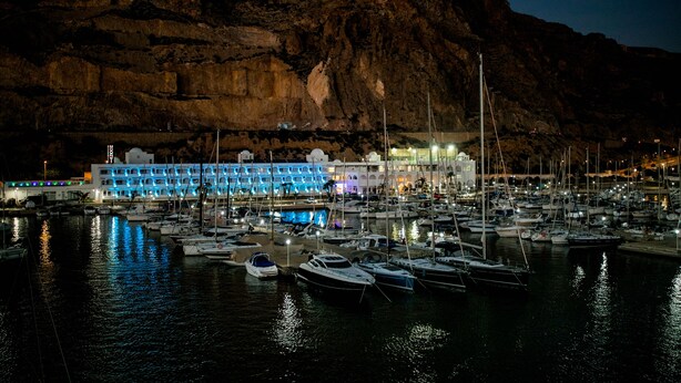 Una marina de noche con barcos amarrados, un edificio iluminado con luces azules y una montaña al fondo.