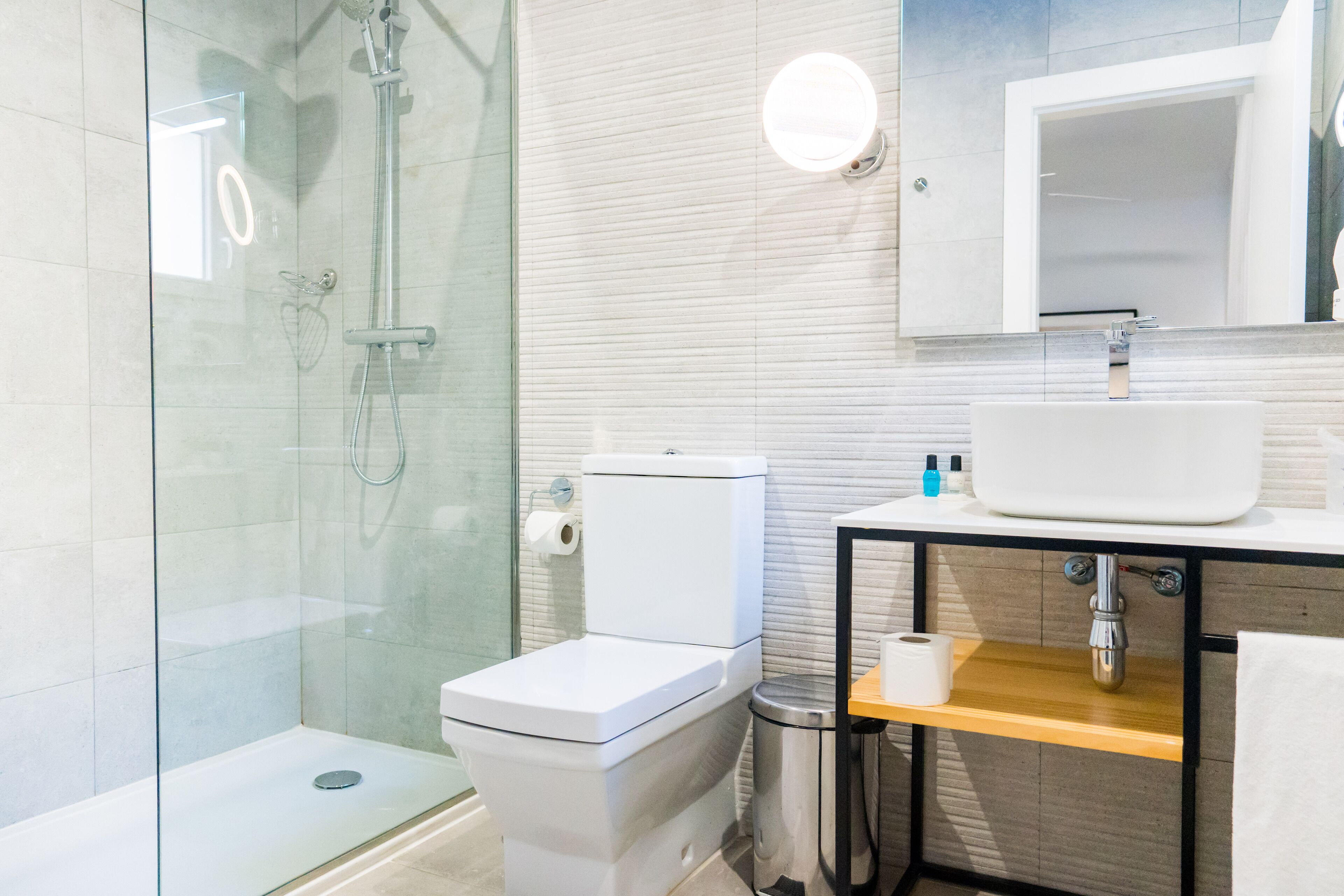 Une salle de bain moderne avec une douche à parois de verre, des toilettes blanches, un lavabo avec un miroir au-dessus et une étagère en bois.