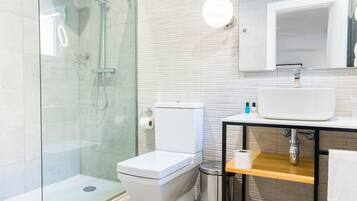 Une salle de bain moderne avec une douche à parois de verre, des toilettes blanches, un lavabo avec un miroir au-dessus et une étagère en bois.