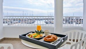 Bandeja de desayuno con croissant, fruta, café y jugo de naranja en un balcón con vista a un puerto deportivo.