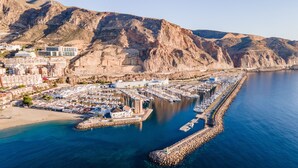 Una marina costera con barcos amarrados, un faro y una montaña rocosa al fondo.