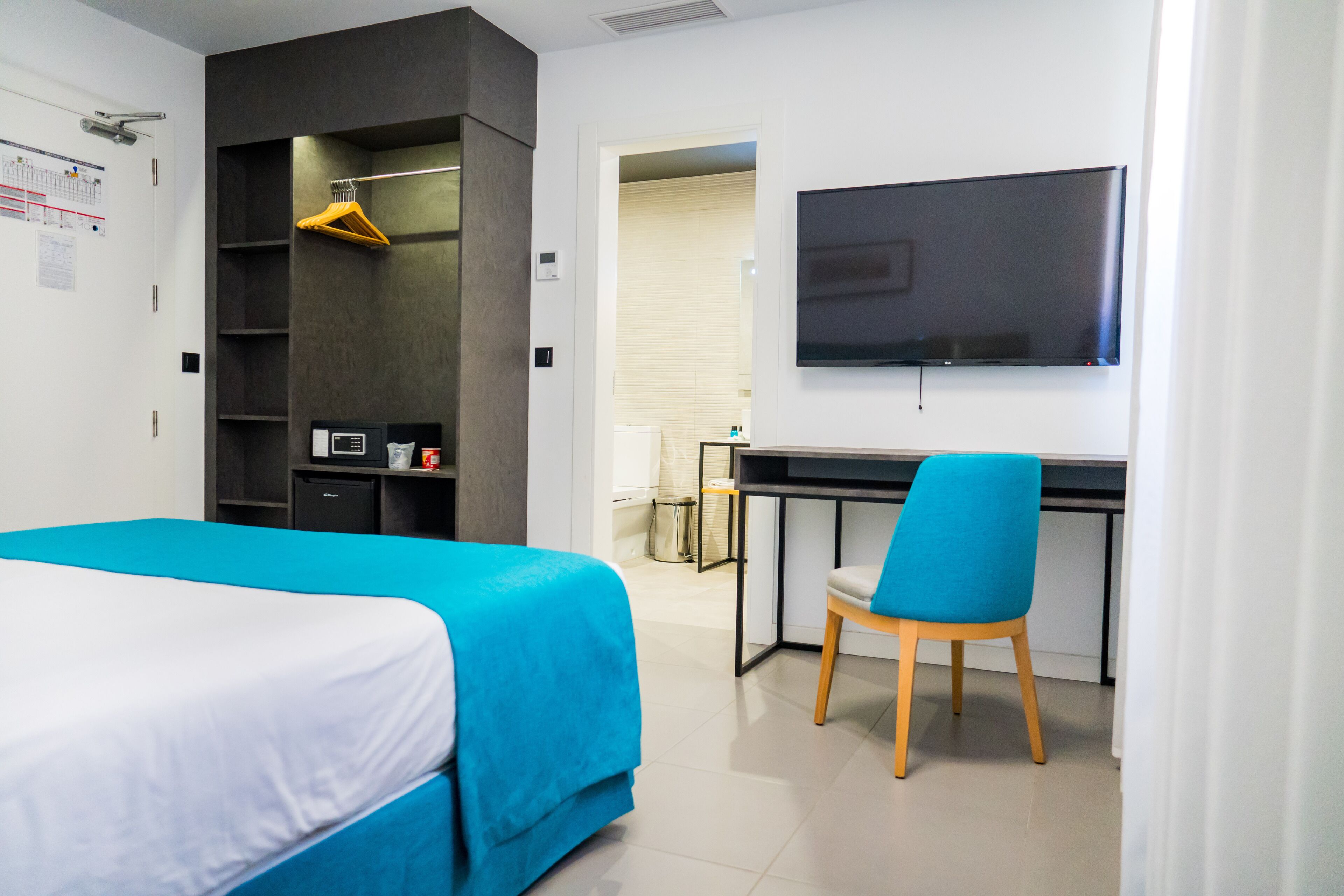A modern hotel room with a bed, a desk, a chair, a TV, and a wardrobe.
