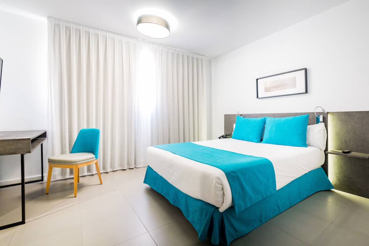 Une chambre d’hôtel avec un lit, une chaise, un bureau et une fenêtre avec des rideaux.