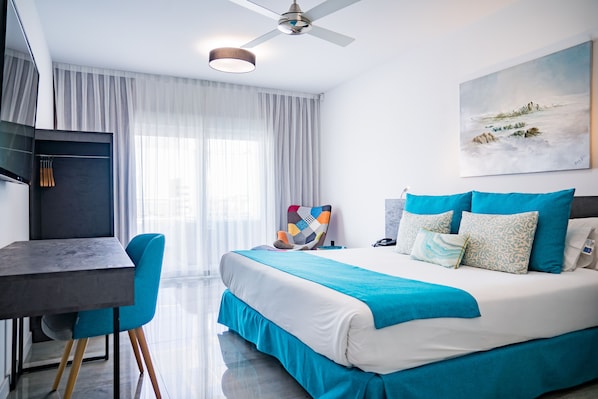 A modern hotel room with a bed, a desk, and a chair. There is a ceiling fan, a painting on the wall, and a window with curtains. - Hotel Moon & SPA (Roquetas de Mar)