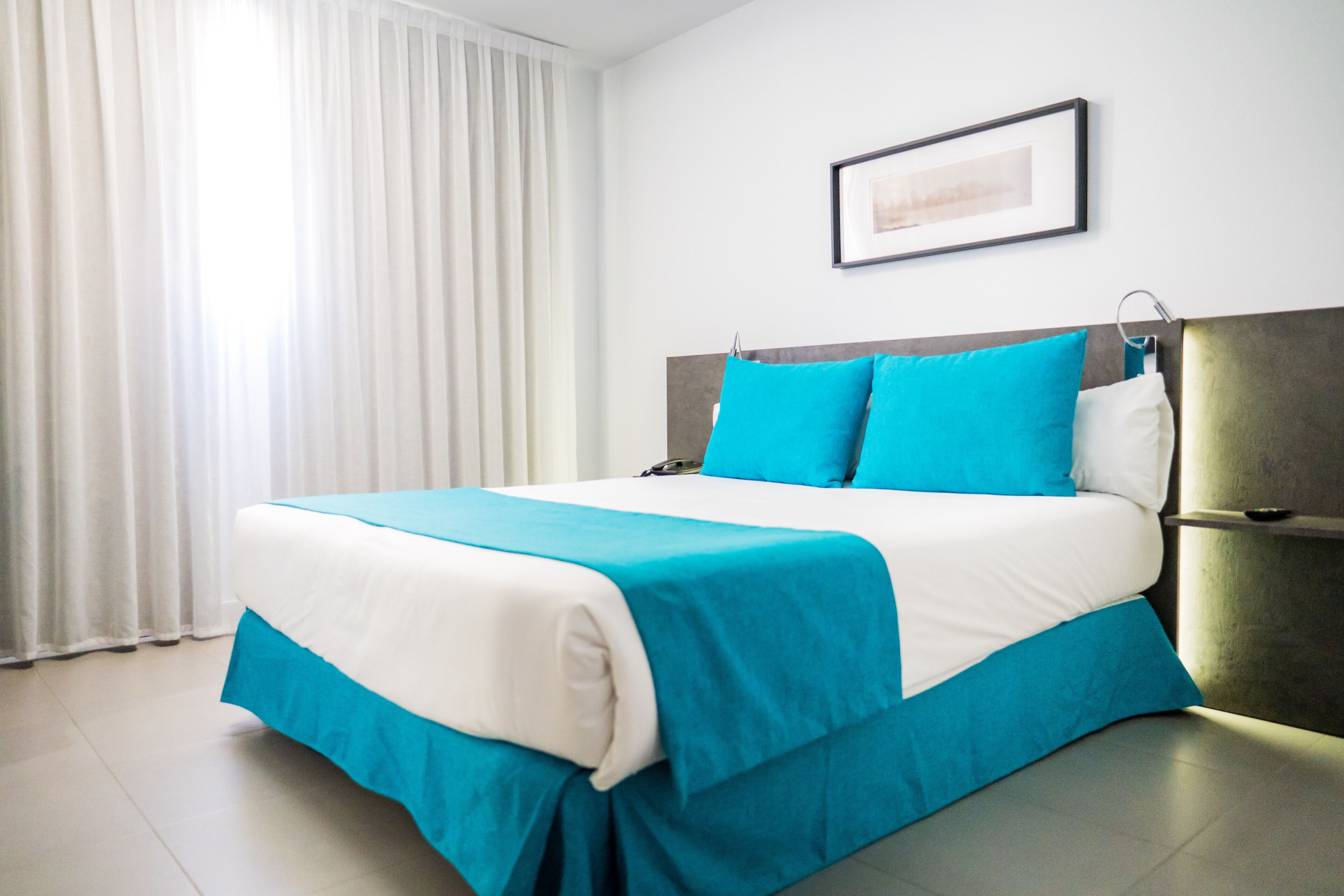 Una habitación de hotel con una cama, un cuadro en la pared y un ventanal con cortinas.