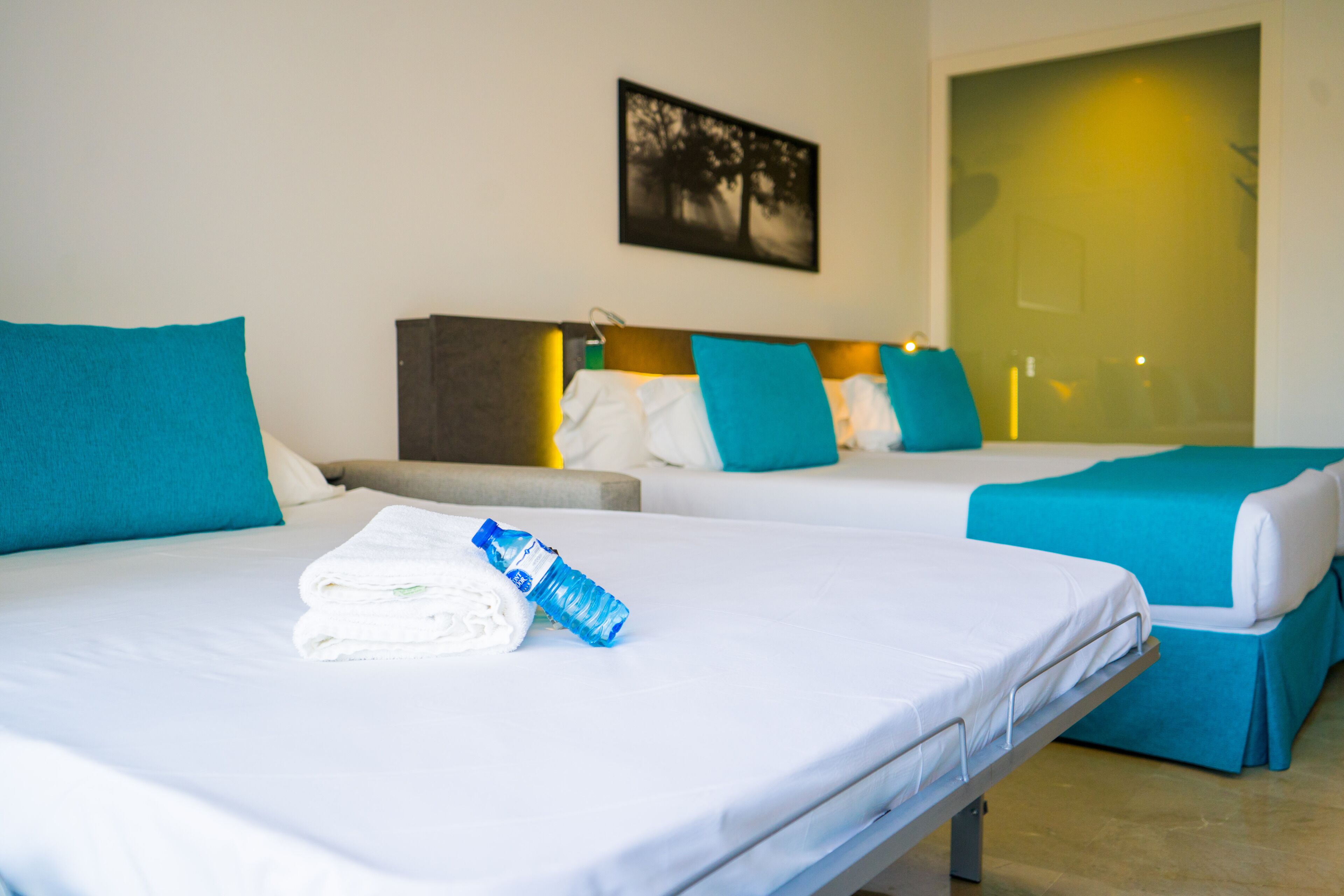 Habitación de hotel con dos camas, cada una con cubrecama azul y blanco, una toalla doblada y una botella de agua sobre la primera cama.