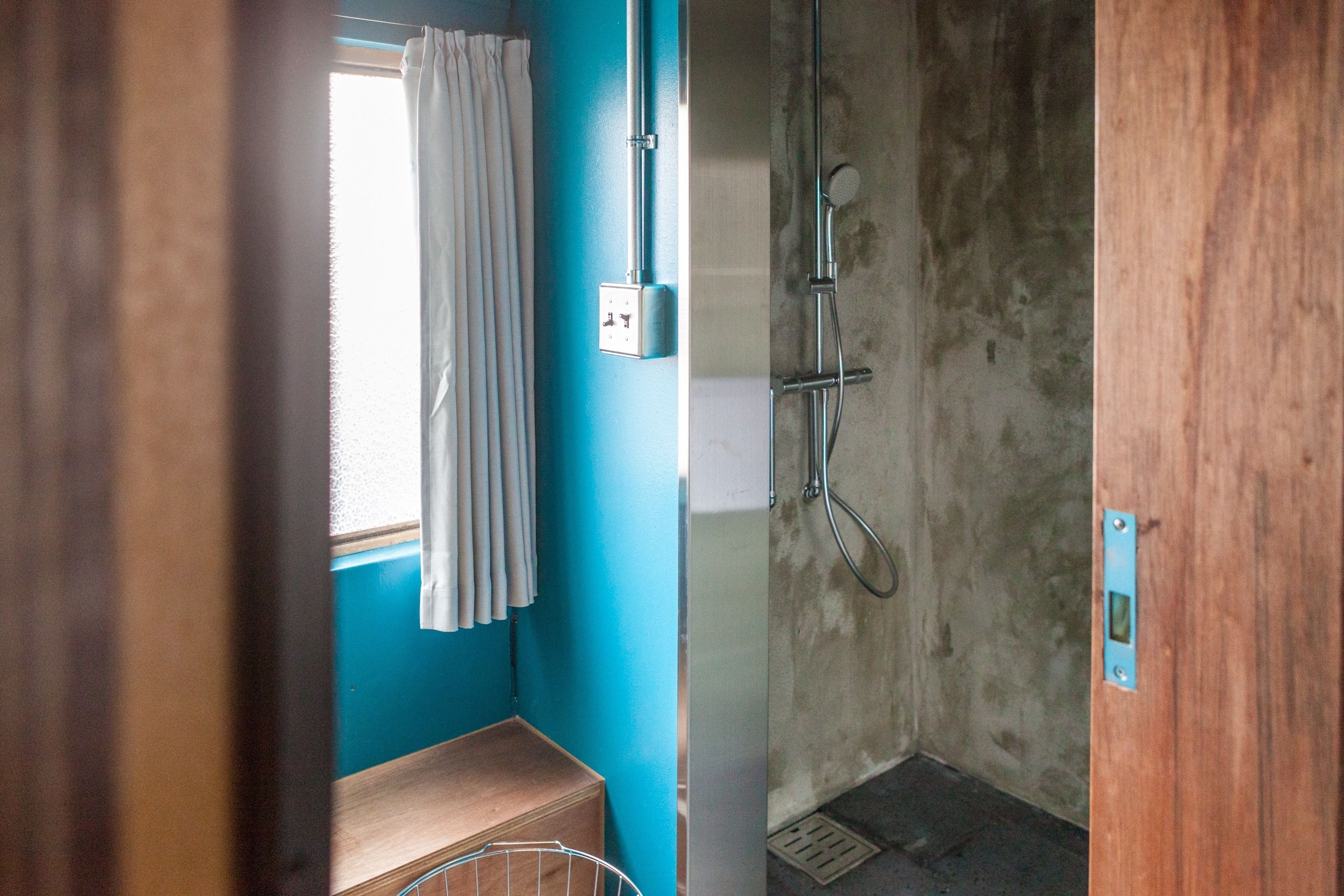 A small, narrow bathroom with a shower, a window with curtains, and a wooden bench.