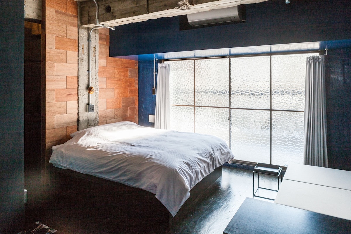A bedroom with a bed, a window with curtains, a wooden wall, and a small table.