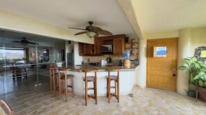 A kitchen with wooden cabinets, a central island with stools, a ceiling fan, and a view through a glass door.