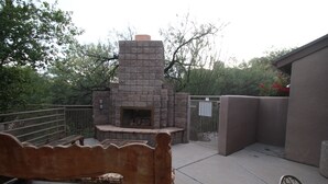Une cheminée en pierre avec un banc devant, le tout entouré d’une terrasse délimitée par une clôture.