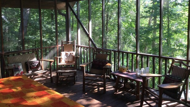 Un espace extérieur aménagé, abrité et doté de mobilier en bois et d’une table, entouré de grandes fenêtres offrant une vue sur une zone boisée.