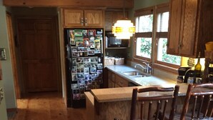 Une cuisine avec des meubles de rangement en bois, un réfrigérateur recouvert de photos, une table à manger en bois avec des chaises, et une fenêtre avec des stores.