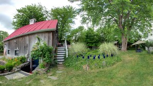 A rustic wooden barn with a red roof, a stone staircase leading up to a porch, and a well-maintained garden with various plants and a small pond.