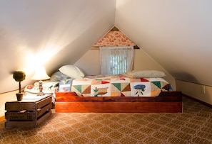 A cozy attic bedroom with a wooden bed, a small table with a lamp, and a window with sheer curtains.
