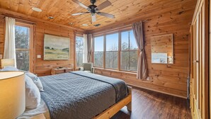 Une chambre dans une cabane en bois, avec un lit, un ventilateur de plafond, un bureau et une fenêtre donnant sur la forêt.