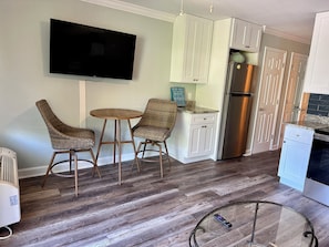 A kitchen with a flat-screen TV, two wicker chairs, a small round table, white cabinets, a stainless steel refrigerator, and a glass-top coffee table.