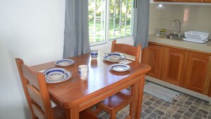 A wooden dining table with blue and white plates, a white mug, and a knife. There is a window with curtains, a kitchen area with wooden cabinets, and a tiled floor.