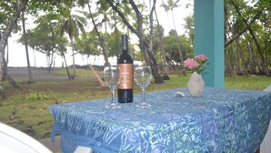 A table with a bottle of wine, two wine glasses, and a vase with flowers, set outdoors with a view of palm trees and a body of water.