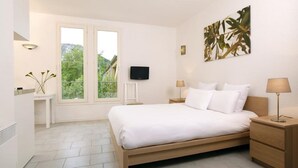 A bedroom with a bed, two bedside tables, a TV, and a window with a view of greenery. - Vacancéole - Le Domaine de Chames (Vallon-Pont-d'Arc)