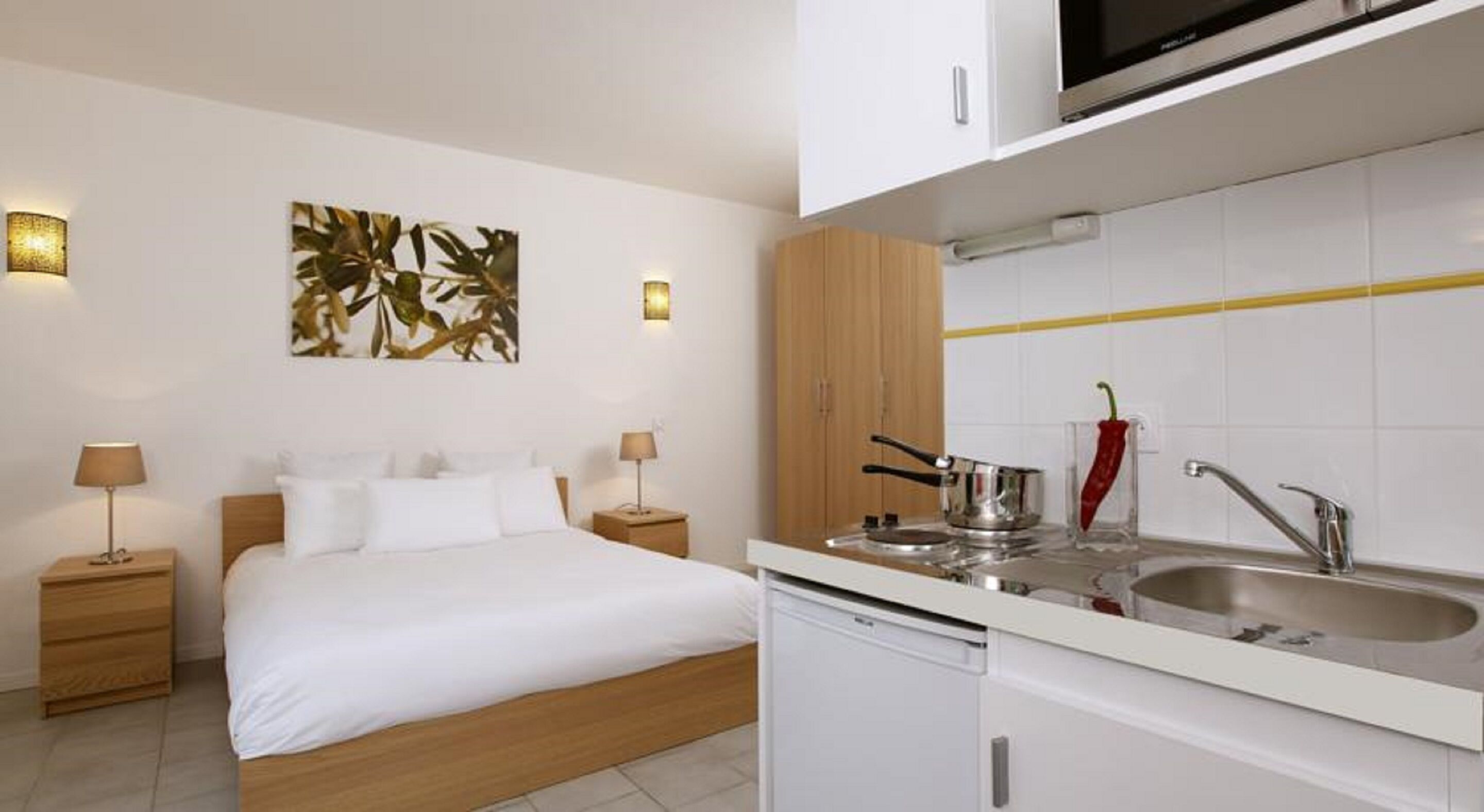 Een hotelkamer met een bed, nachtkastjes, een schilderij en een keuken met een gootsteen, fornuis en een rode chilipeper.