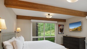 A bedroom with a bed, bedside lamps, a dresser, a television, and a window with a view of trees.