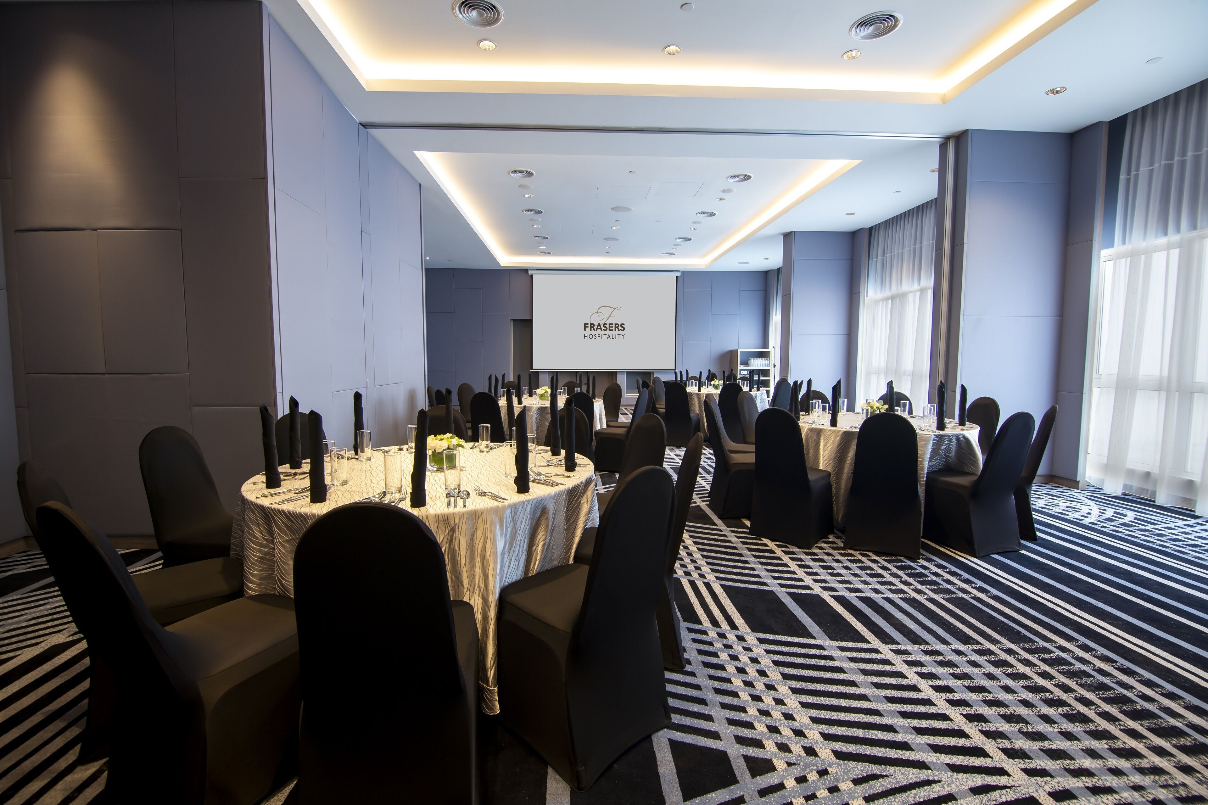 A conference room with round tables, black chairs, and a projector screen displaying "FRASES HOSPITALITY".