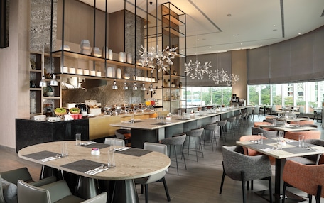 A modern restaurant with a central bar, multiple dining tables, and a large window offering a view of the outdoors.