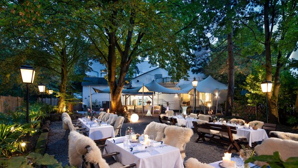 An outdoor dining area with tables set for a meal, surrounded by trees and a building in the background. - Fettehenne Gästehaus (Erkrath)