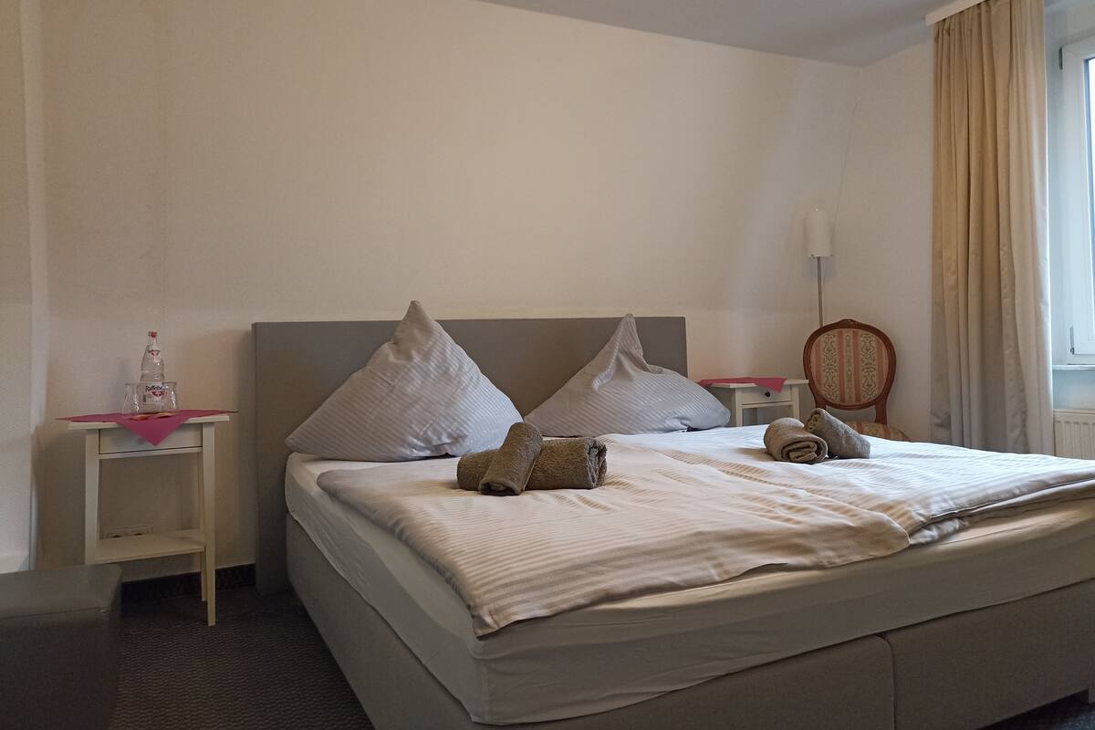 A neatly made bed with pillows, a bedside table with a bottle, and a chair.