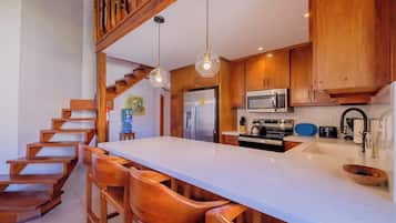 A modern kitchen with wooden cabinetry, a central island with seating, stainless steel appliances, and a staircase leading to an upper level.