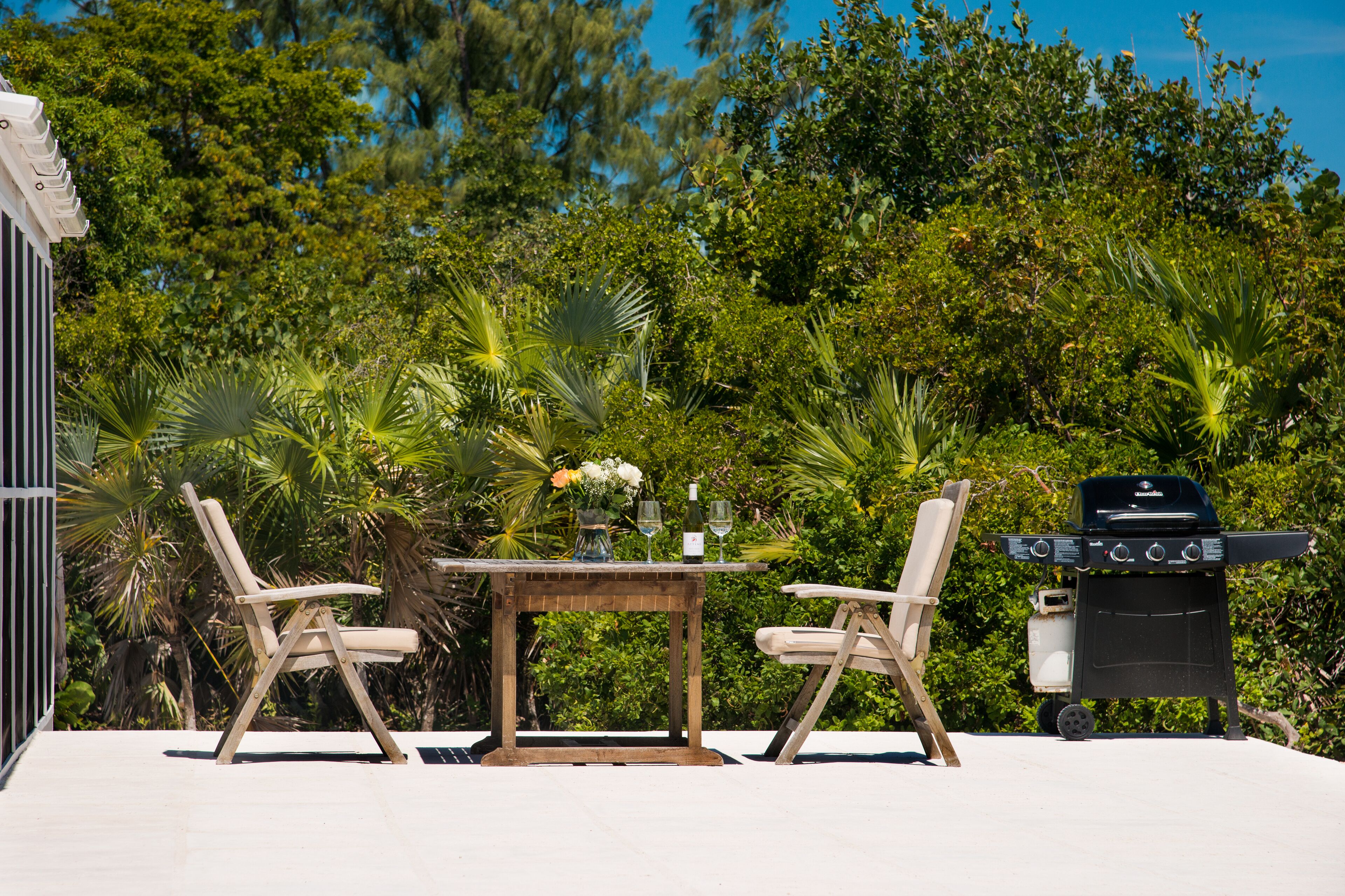 An outdoor patio with a wooden table, two chairs, a grill, and lush greenery in the background.