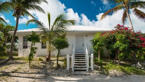 A white building with a front door and steps leading up to it, surrounded by palm trees and flowering bushes. - Romantic Getway Steps from the Beach (Providenciales)