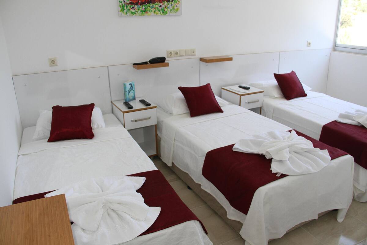 A hotel room with four single beds, each with a white bedspread and a red pillow. There are bedside tables with a remote control and a phone on each. The room has a white wall with a small shelf above the beds.