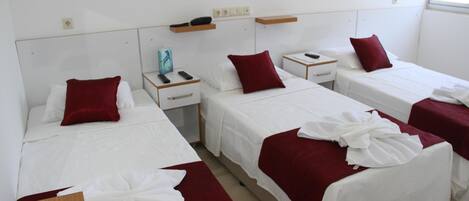 A hotel room with four single beds, each with a white bedspread and a red pillow. There are bedside tables with a remote control and a phone on each. The room has a white wall with a small shelf above the beds.