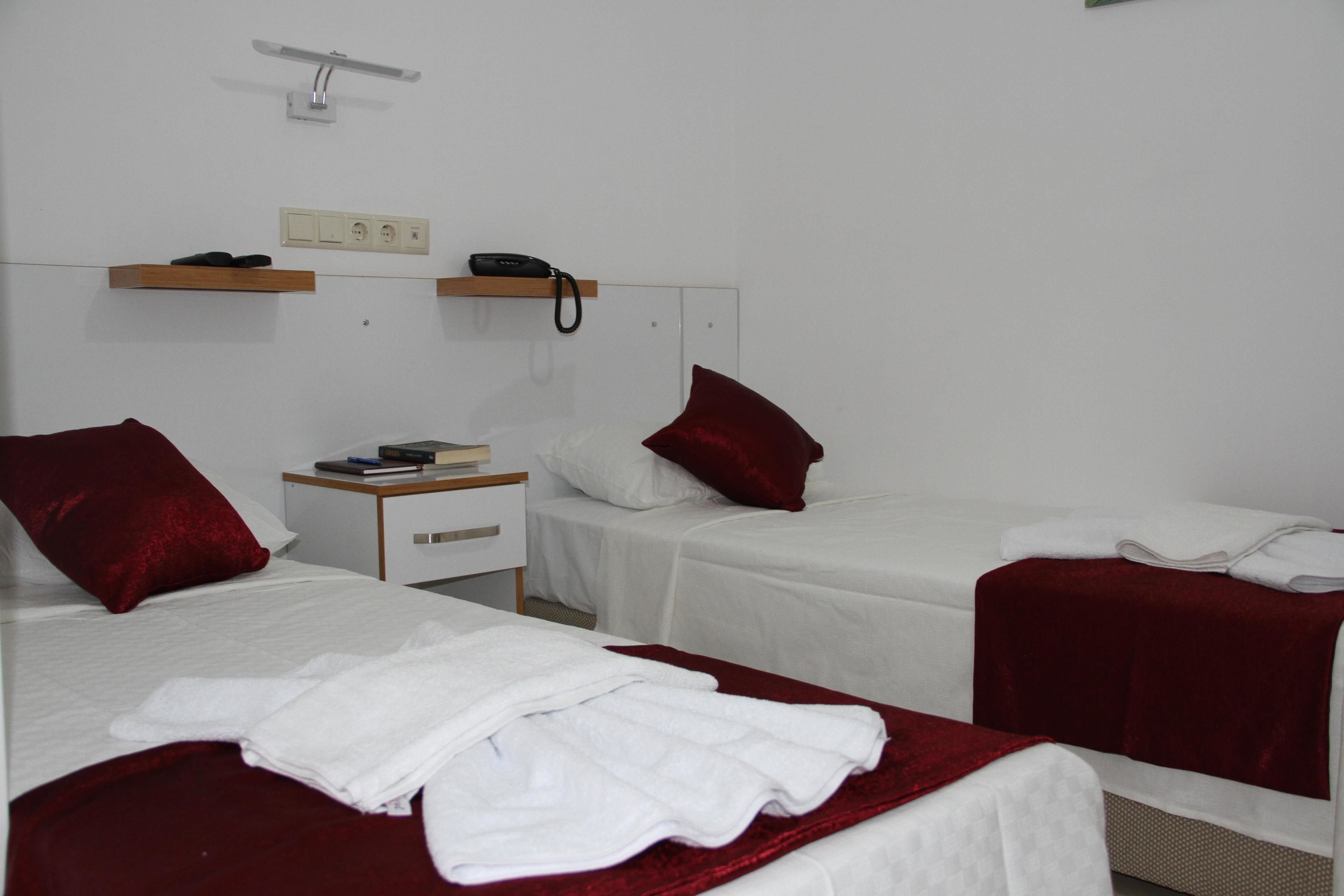 A hotel room with two beds, each with a red pillow and white bedding. There is a bedside table with a book and a phone.