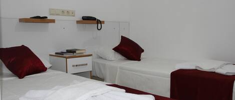 A hotel room with two beds, each with a red pillow and white bedding. There is a bedside table with a book and a phone.